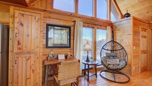 A cozy cabin interior with wooden walls, a large window, a wicker chair, and a small table with a lamp. - All Ya Need 4 Bedroom Cabin (Sevierville)