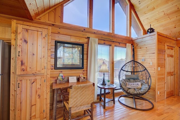 A cozy cabin interior with wooden walls, a large window, a wicker chair, and a small table with a lamp. - All Ya Need 4 Bedroom Cabin (Sevierville)