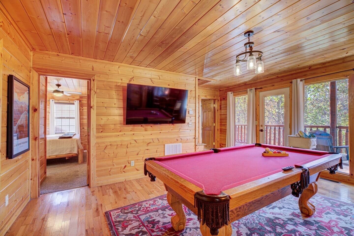 A wooden interior with a pool table, a television mounted on the wall, and a door leading to another room.