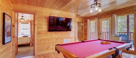 A wooden interior with a pool table, a television mounted on the wall, and a door leading to another room.