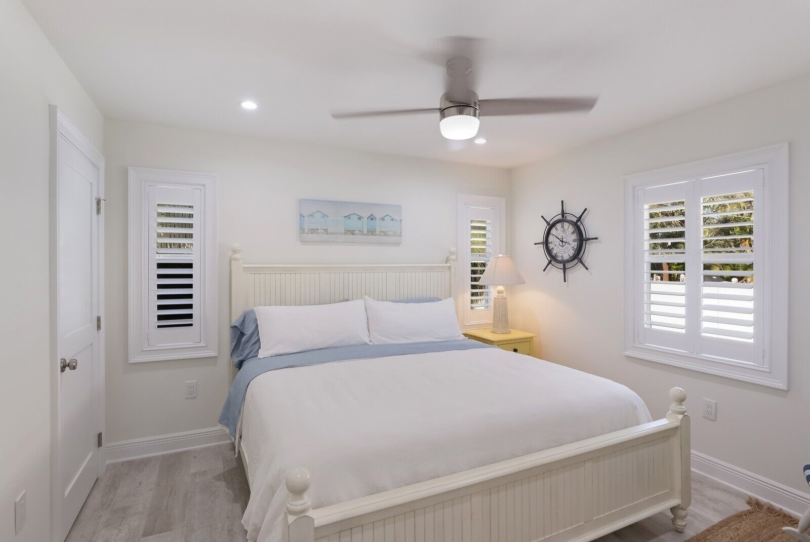 Une chambre à coucher avec un lit blanc, un ventilateur de plafond, une horloge murale et une fenêtre avec des volets.