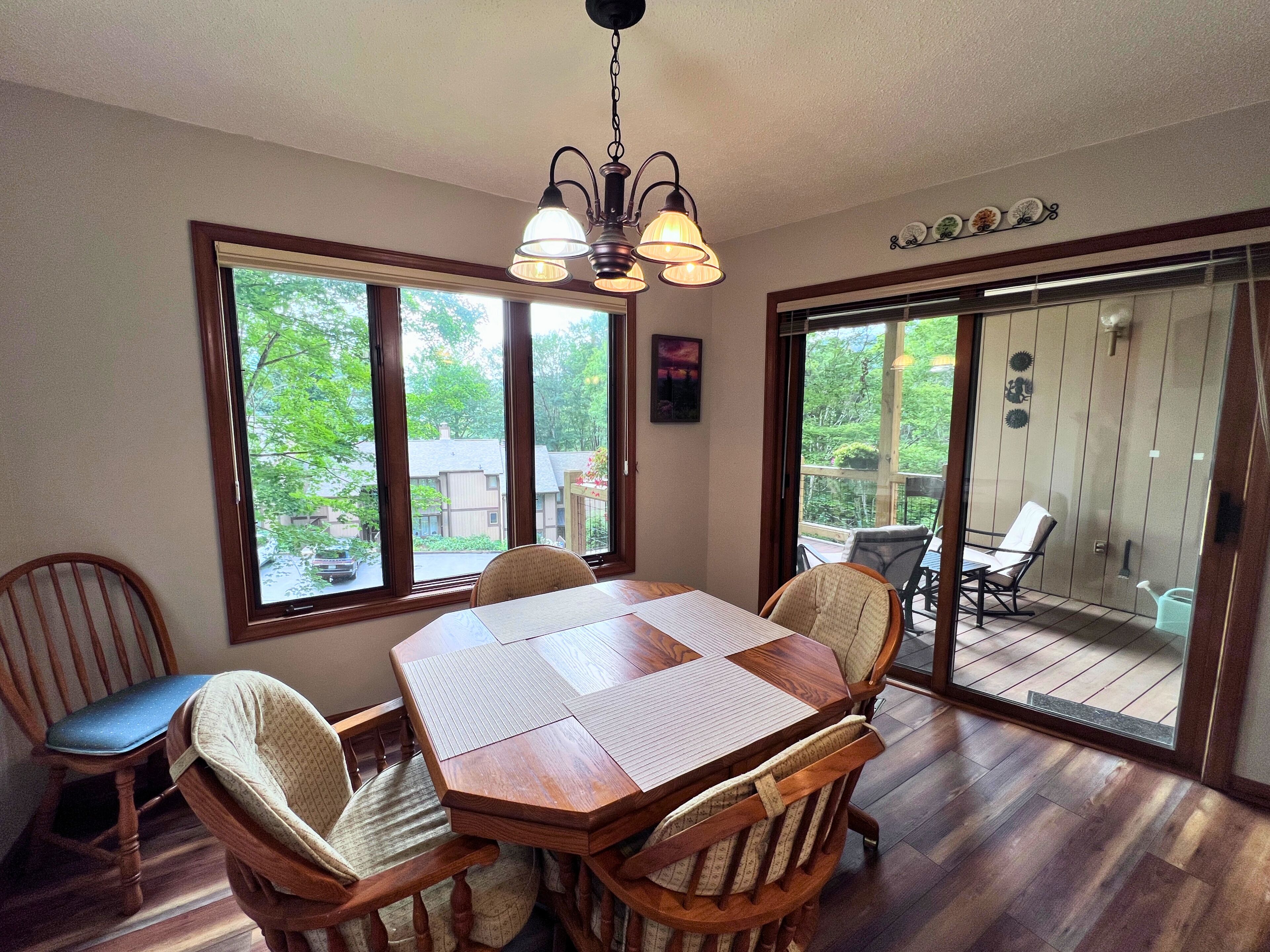 A dining area with a wooden table and chairs, large windows, and a view of the outdoors.