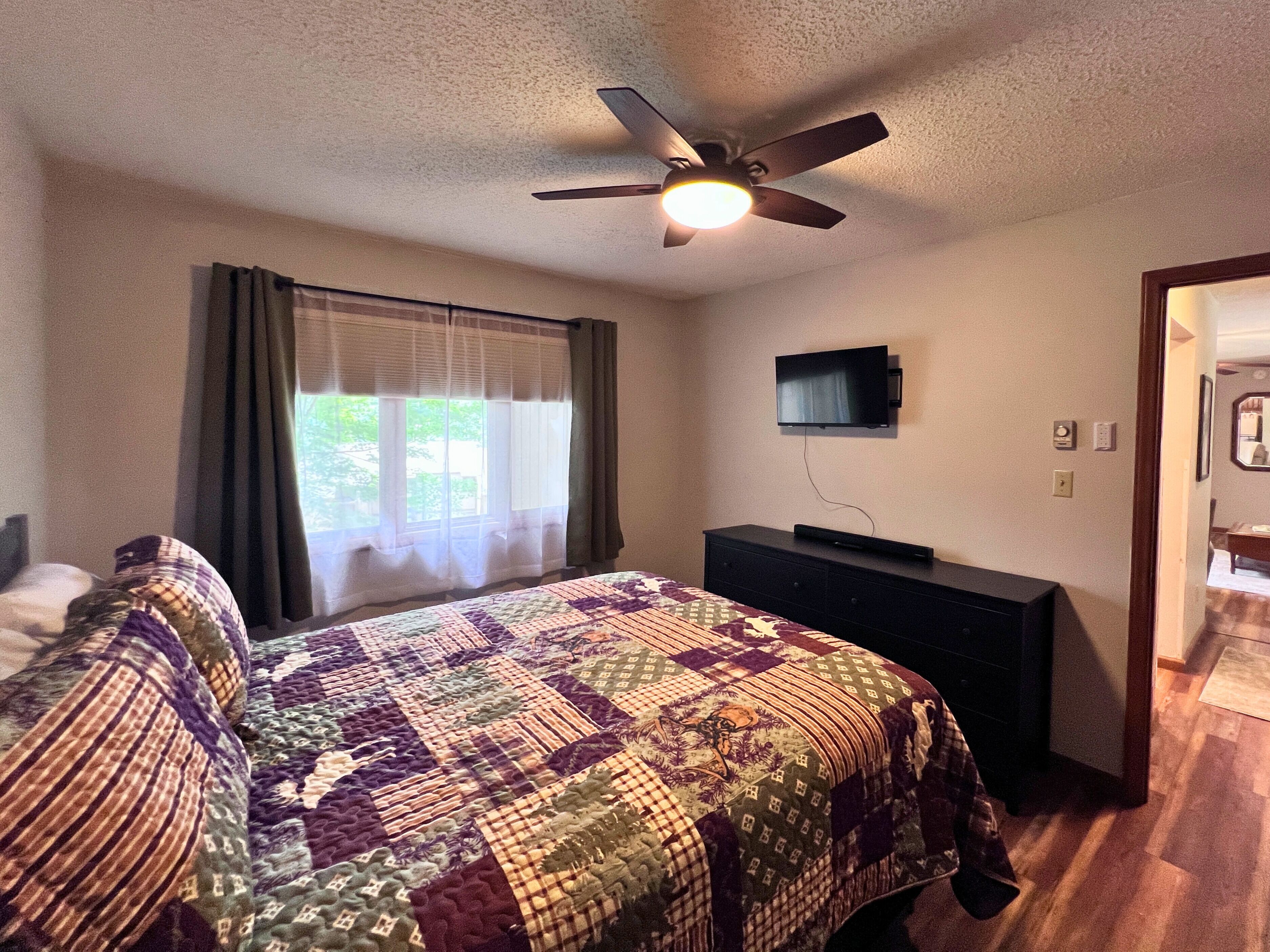 A bedroom with a bed, a dresser, a ceiling fan, a TV, and a window with curtains.