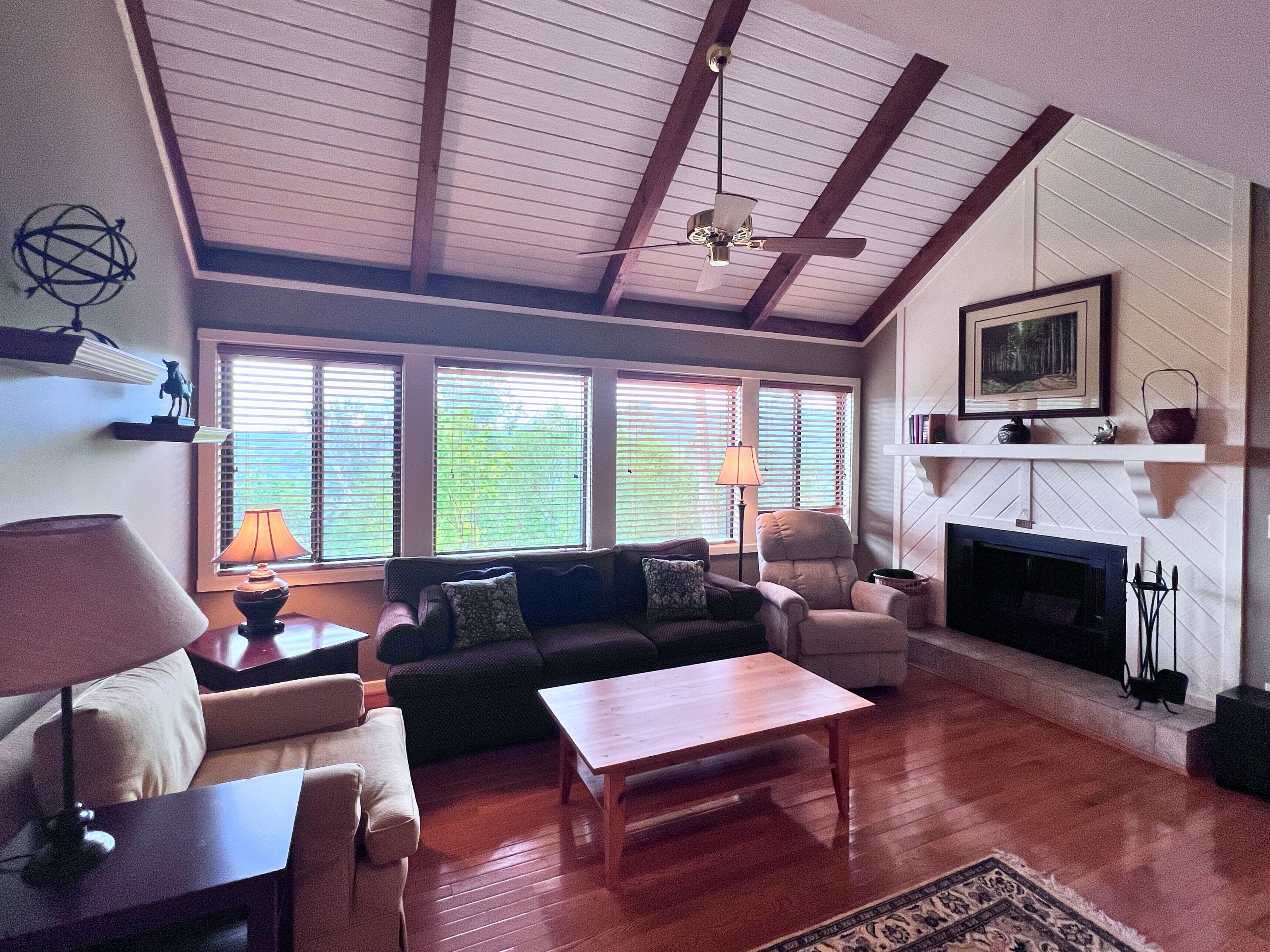 A spacious living room with a fireplace, wooden flooring, and a high ceiling with exposed beams.
