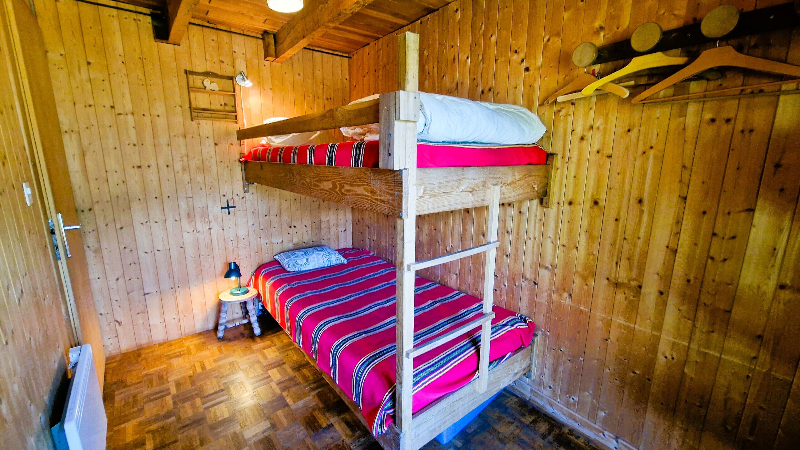 Quarto de cabana de madeira com beliche, uma mesinha com abajur e uma estante com objetos pessoais.
