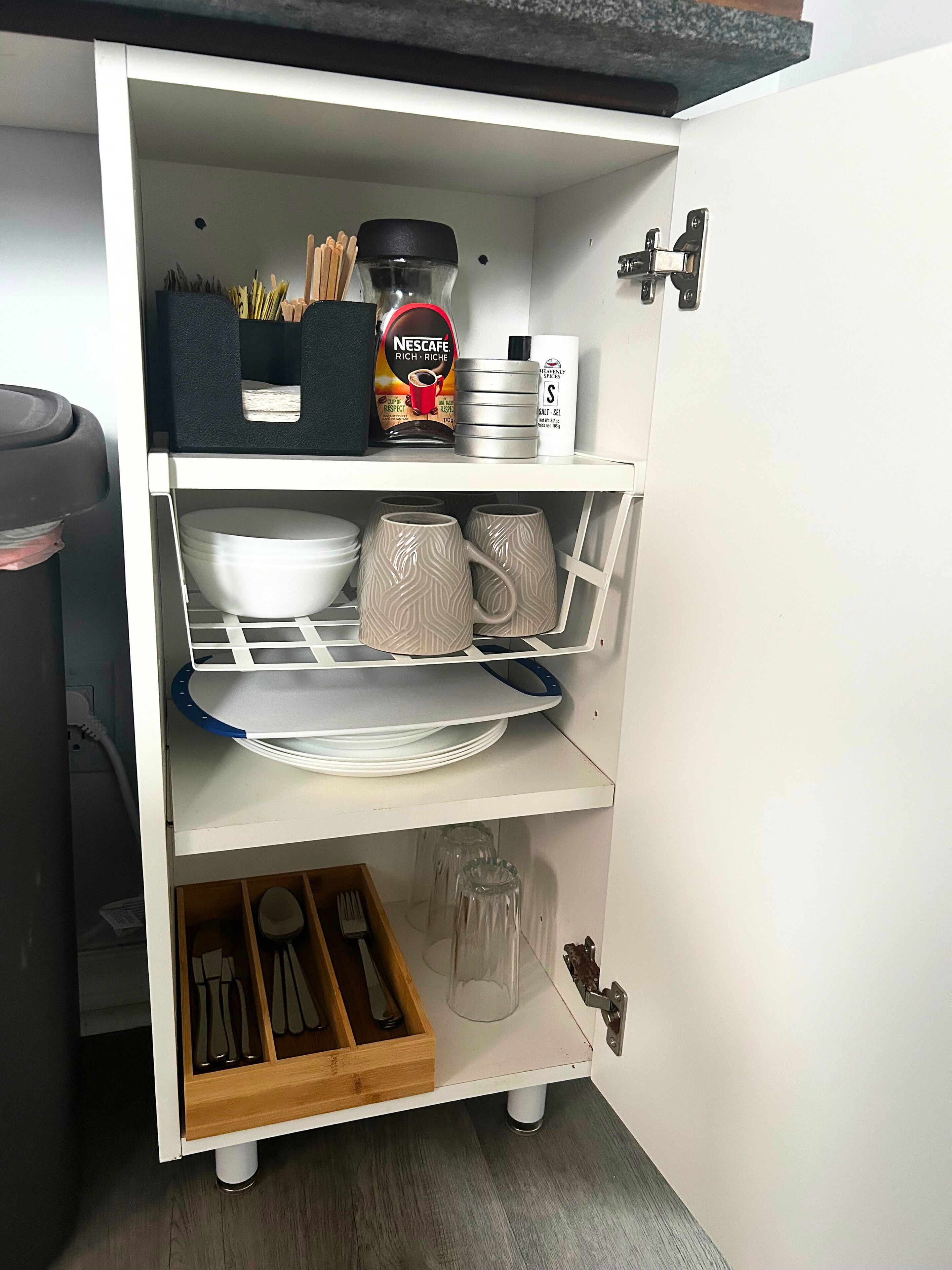 A white kitchen cabinet with shelves containing dishes, mugs, a coffee maker, and utensils.