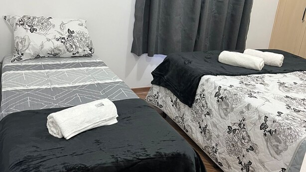 A room with two beds, each with a black and white floral bedspread, a grey curtain, and a wooden floor. - Apartment in front of Colinas shopping mall (Jardim das Colinas São José dos Campos)