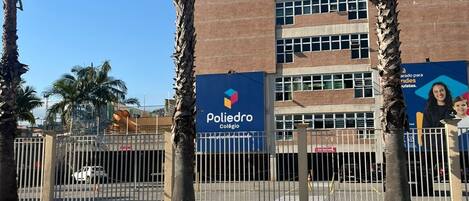 A building with a blue sign reading "Poliedro Colégio" and a fenced area with palm trees.