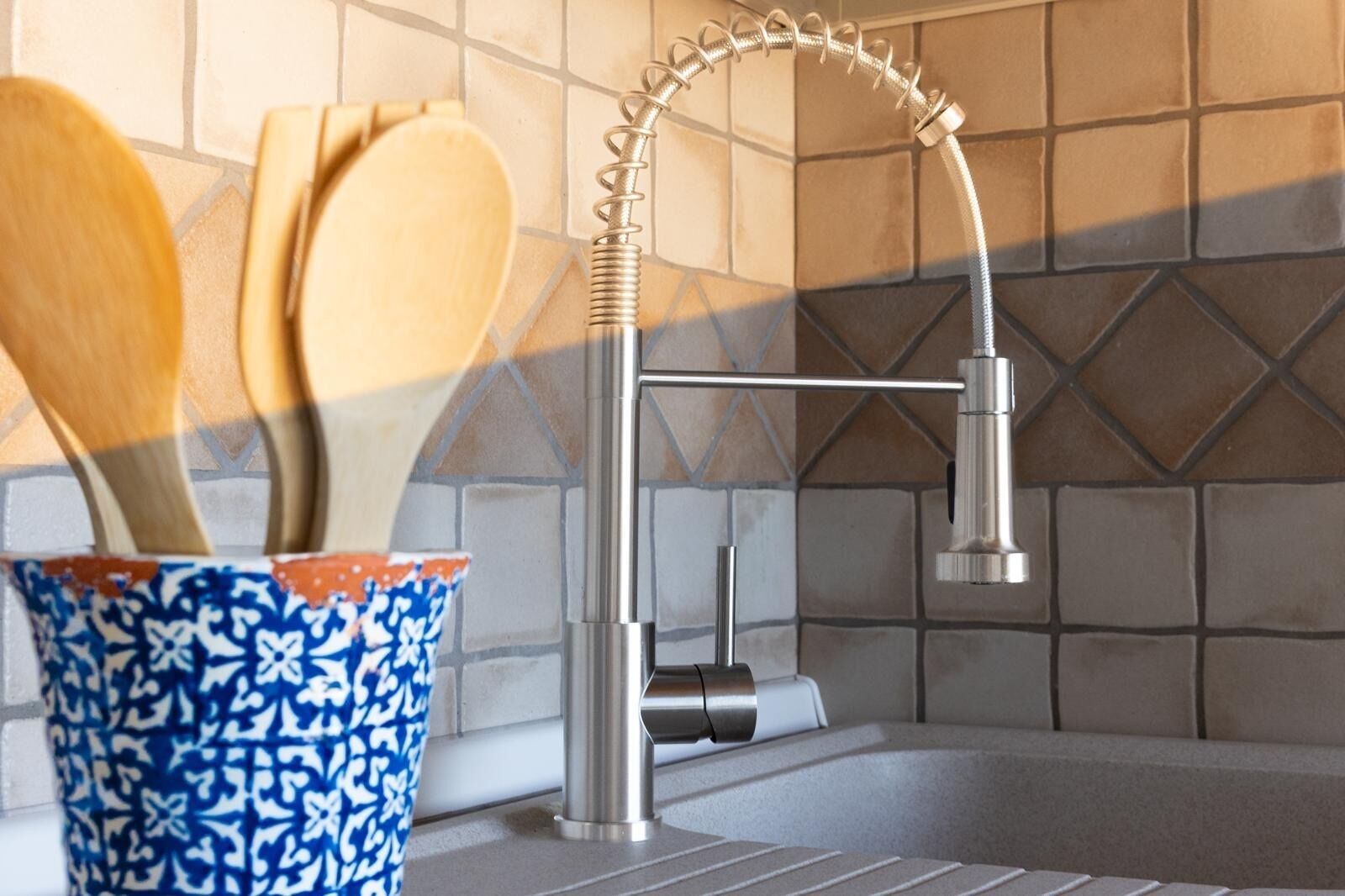 A modern kitchen with a stainless steel faucet, a wooden spoon in a patterned bowl, and a tiled backsplash.