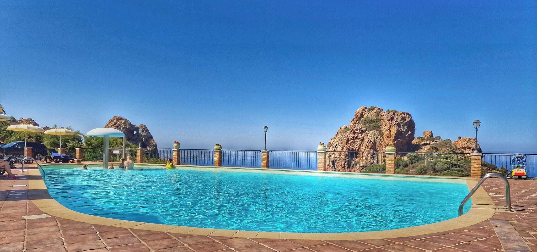 A clear blue pool with a tiled deck, umbrellas, and a view of rocky cliffs.