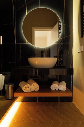 A modern bathroom with a large oval mirror, a white vessel sink, and neatly rolled towels on a wooden shelf. - GV Suite Collection (Rome)