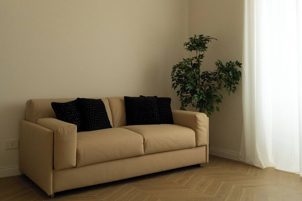 A room with a beige sofa, a potted plant, and a white curtain.