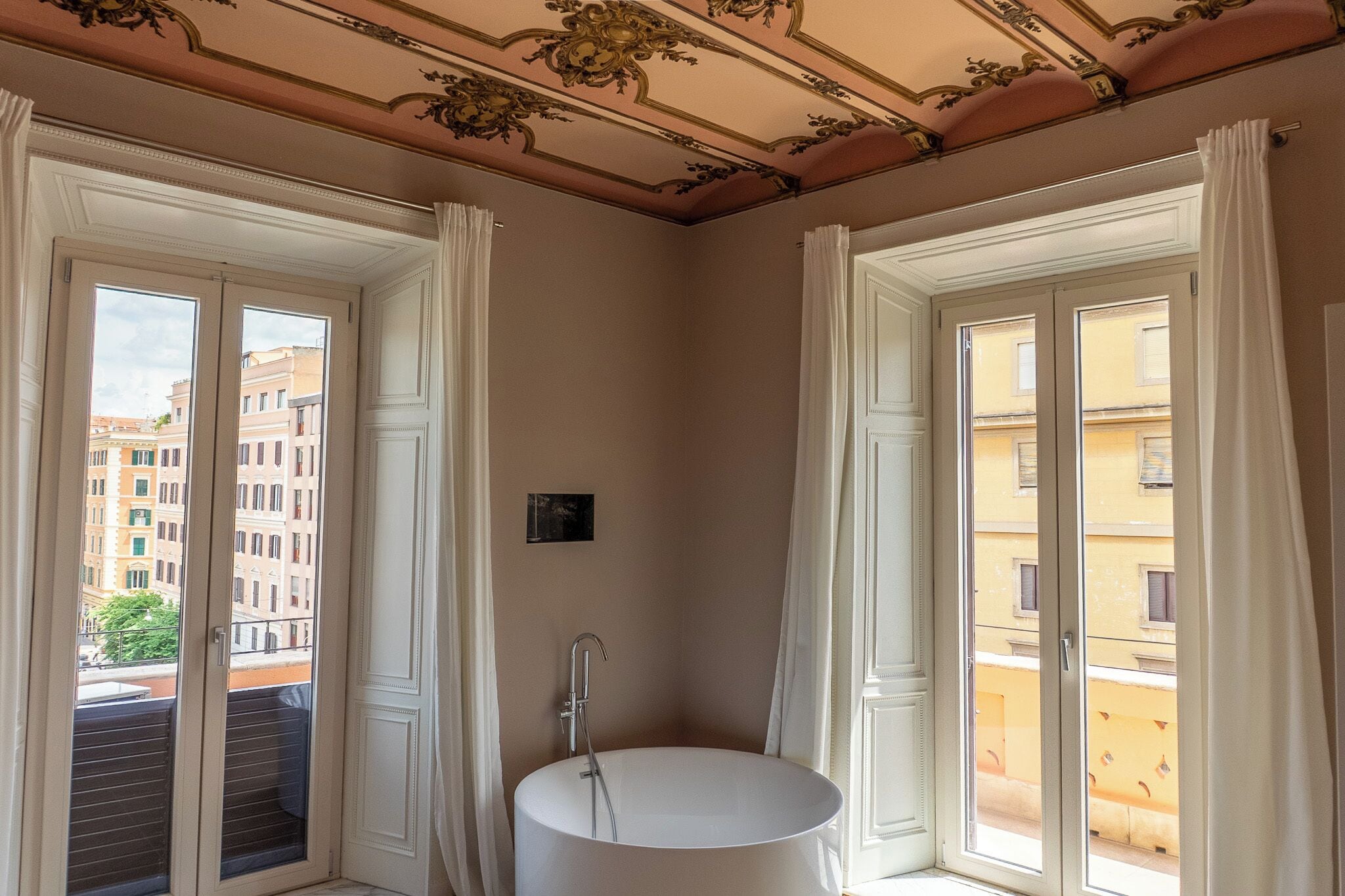A corner bathroom with a freestanding bathtub, large windows with curtains, and a view of buildings outside.