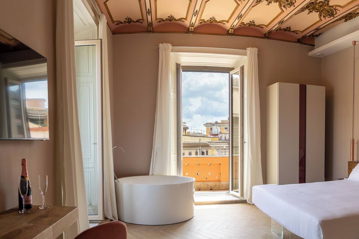 A hotel room with a bed, a seating area, a wardrobe, and a view of buildings through a window.