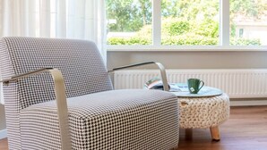 A houndstooth patterned armchair with a metal frame, positioned next to a round ottoman with a knitted base, in a room with large windows and a radiator. - Rustic and spacious vacation home on a small vacation park in the middle of Drenthe (Schoonloo)