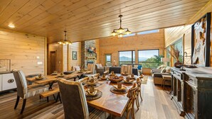 A spacious dining area with wooden tables and chairs, a stone fireplace, and large windows offering a view of the outdoors. - Swim in the Clouds - Luxe 6BR Cabin w/ Views & Pool (Pigeon Forge)