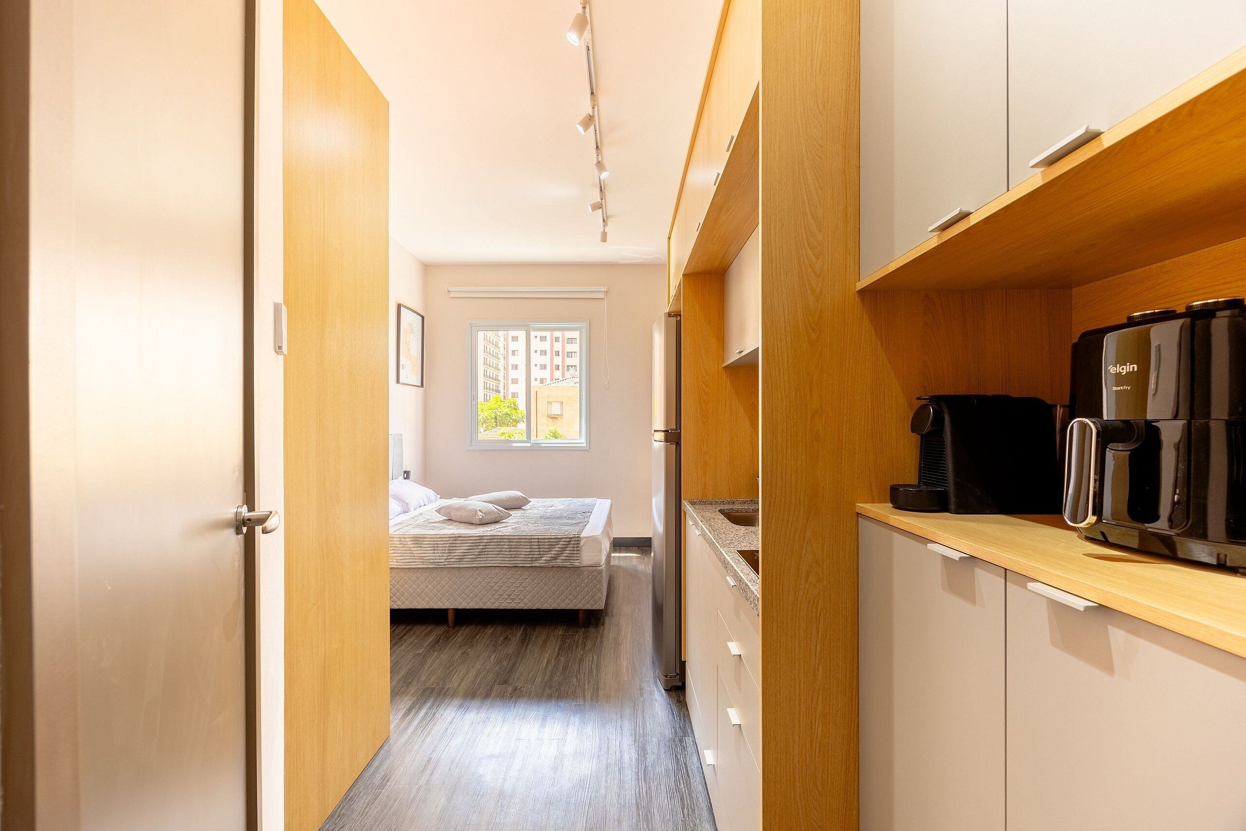A compact hotel room with a kitchenette, a bed with pillows, a window with blinds, and a door leading to another room.