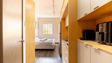 A compact hotel room with a kitchenette, a bed with pillows, a window with blinds, and a door leading to another room.