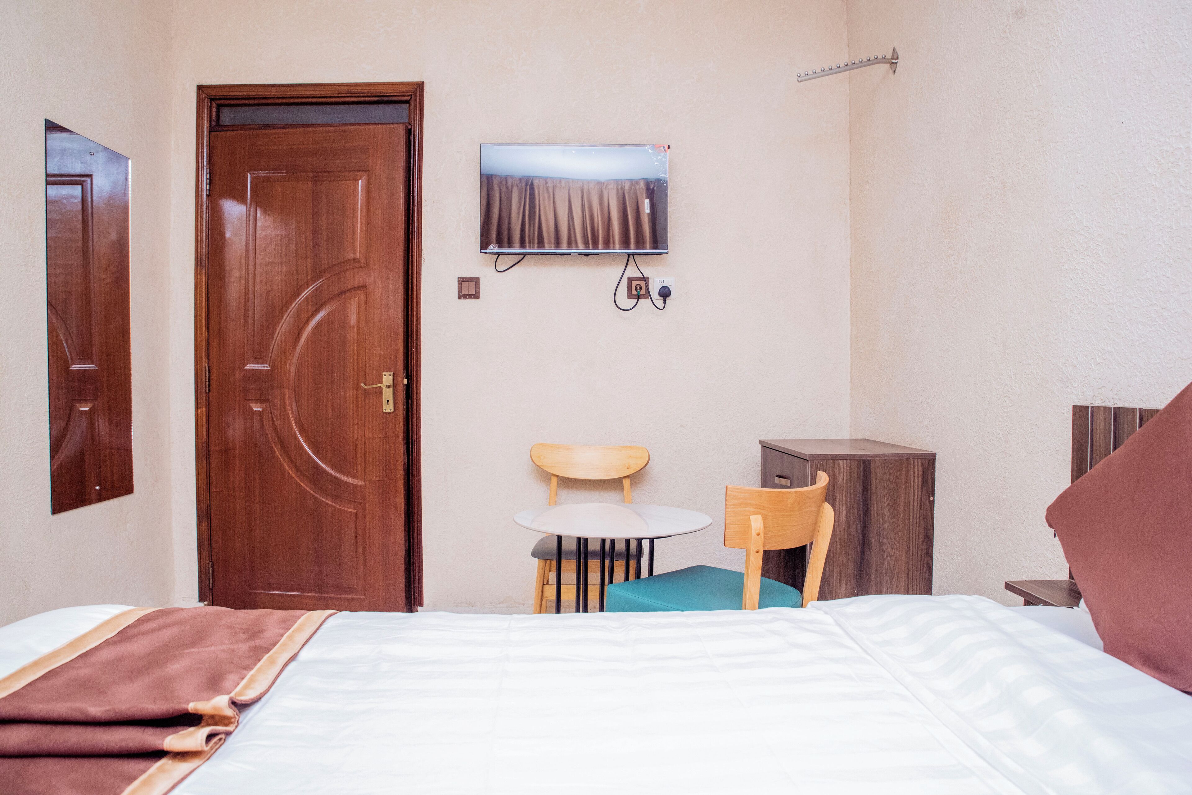 Una habitación de hotel con cama, una puerta de madera, una mesita redonda con una silla, un televisor montado en la pared y un mueble pequeño.