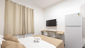 A hotel room with a bed, a desk with a chair, a flat-screen TV mounted on the wall, and a white refrigerator.