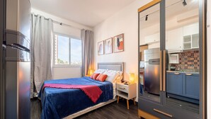 A bedroom with a bed, bedside table, and a view of the kitchen through an open door. - Modern studio in São Caetano Ap709 (São Caetano do Sul)