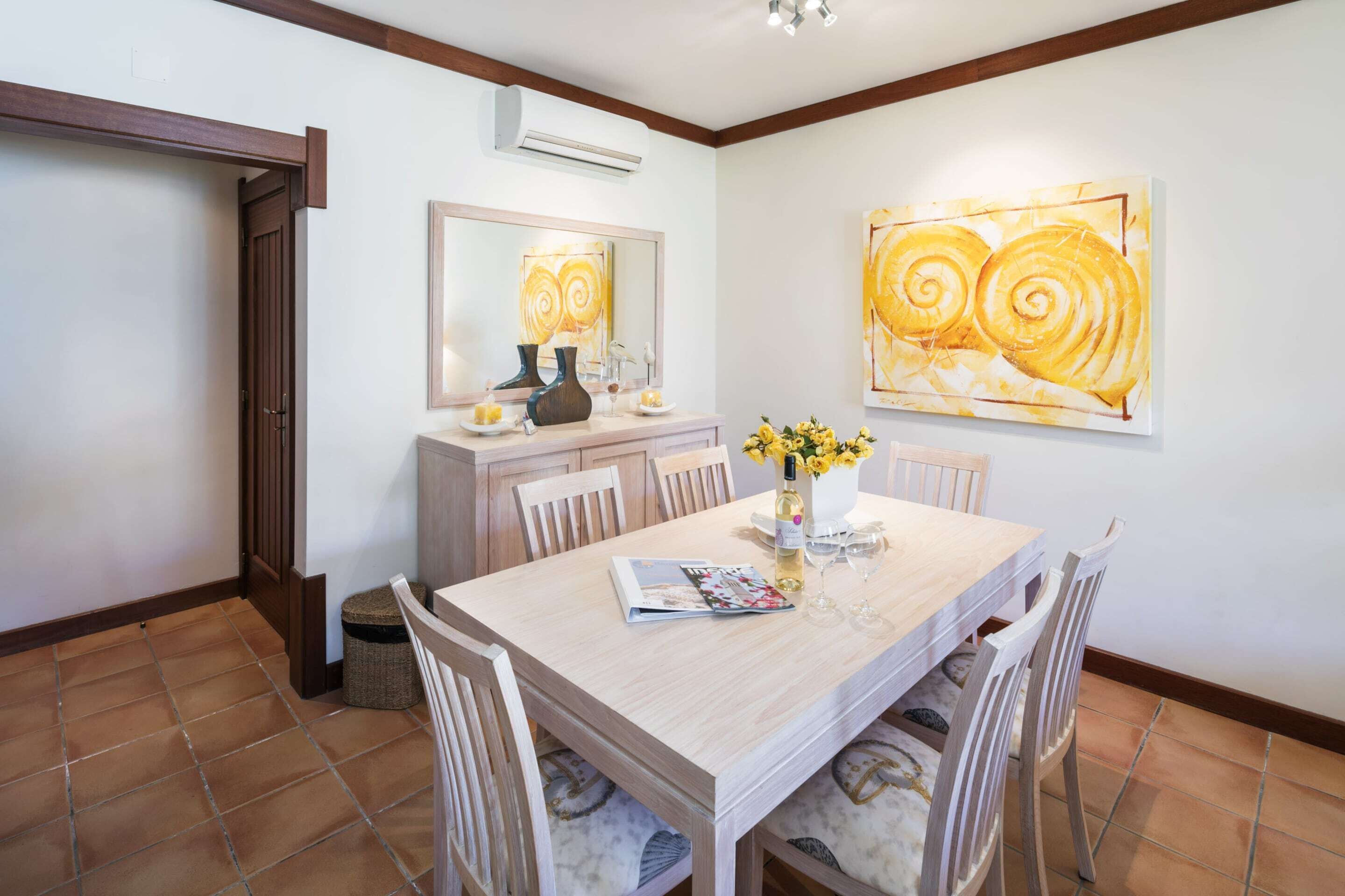 A dining area with a wooden table, chairs, a vase with flowers, a magazine, and a painting on the wall.