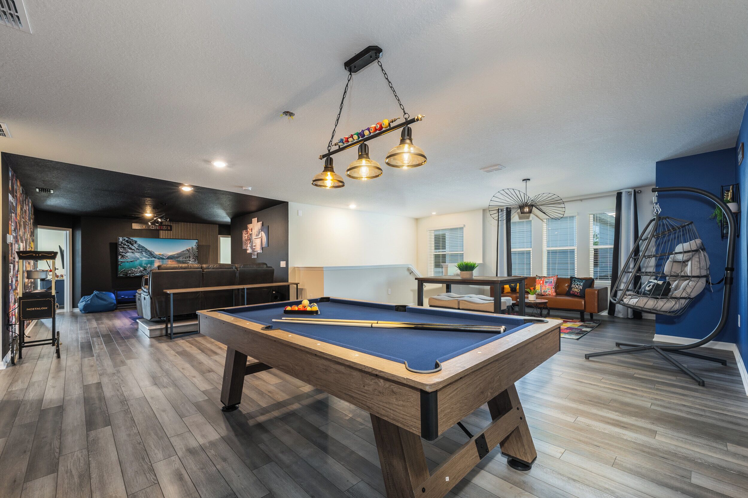 A pool table in a modern living room with a hanging light fixture, a bean bag chair, and a large window.