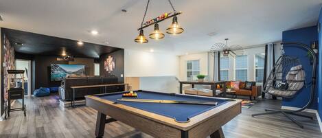 A pool table in a modern living room with a hanging light fixture, a bean bag chair, and a large window.
