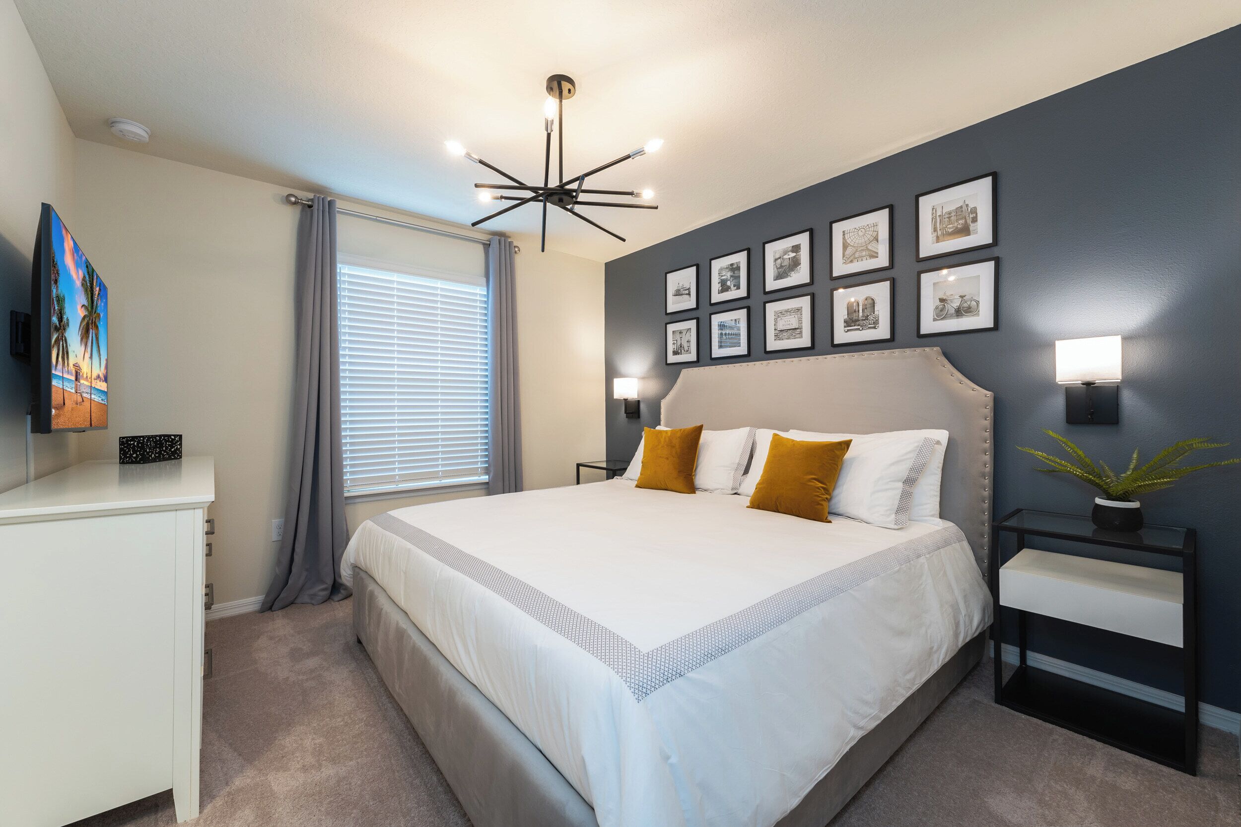 A bedroom with a large bed, a bedside table, a nightstand, a ceiling light fixture, a window with curtains, and a wall with framed pictures.