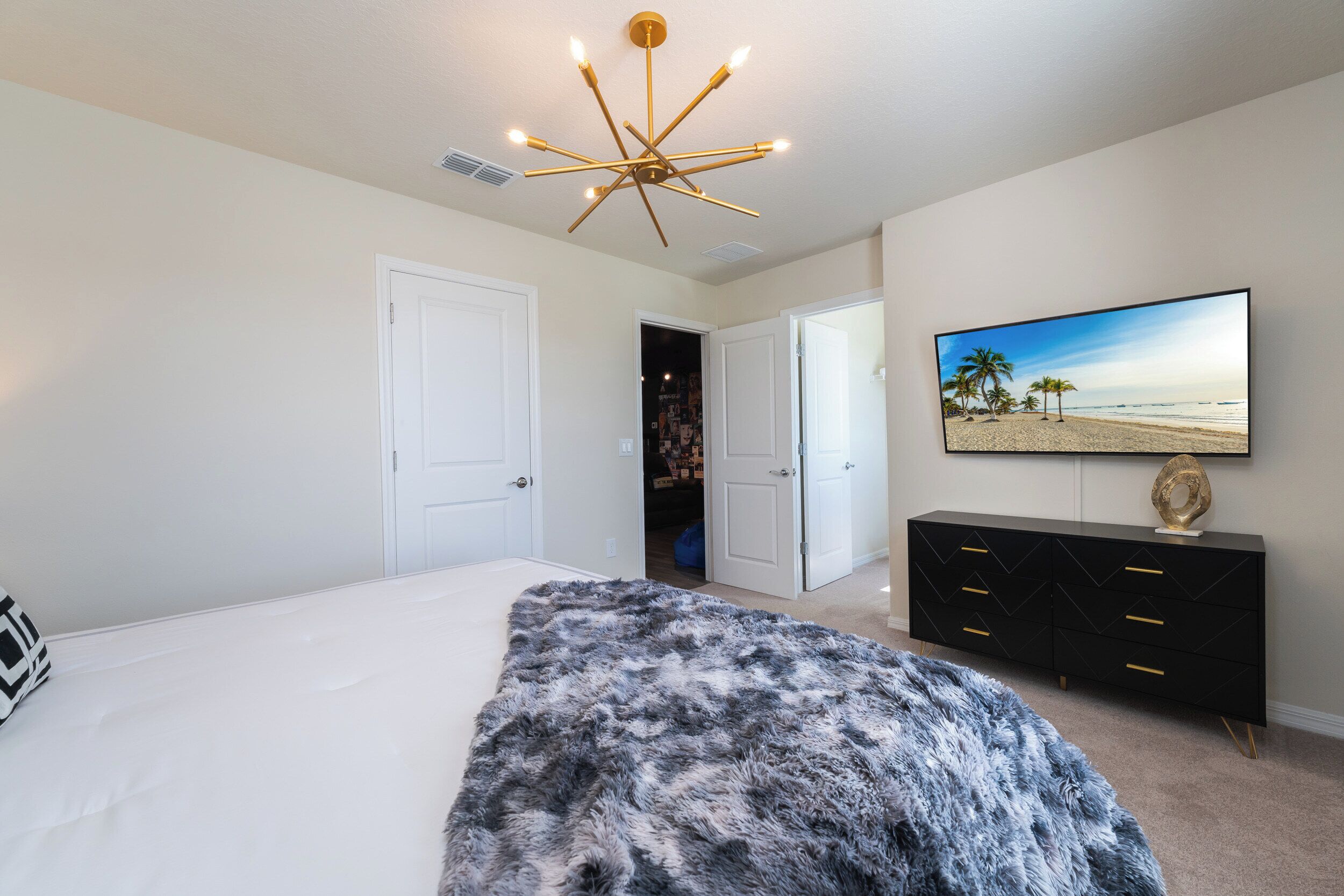 A bedroom with a bed, a fluffy rug, a black dresser, a TV on the wall, and a door leading to another room.