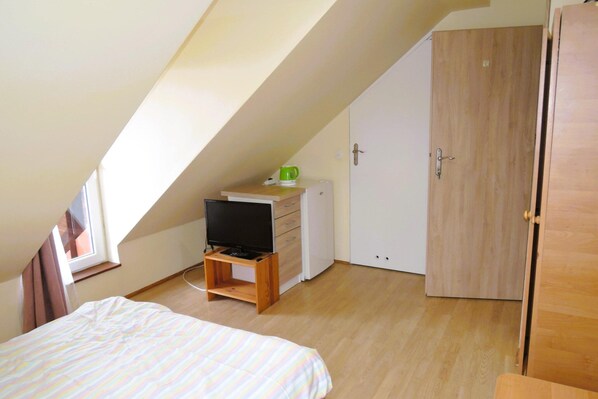 Una habitación pequeña con techo inclinado, una cama, un televisor sobre un soporte de madera, un armario y una puerta.
