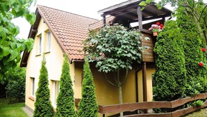 Une maison avec un toit en tuiles, un porche en bois et un jardin où les haies sont soigneusement taillées, le tout délimité par une clôture en bois.
