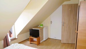 Une petite pièce bien éclairée, avec un plafond en pente, un lit, un téléviseur posé sur un meuble en bois, une armoire et un petit placard.
