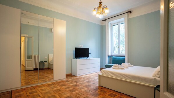 Een slaapkamer met een grote spiegel, een televisie op een dressoir, een bed met een blauw hoofdeinde en een raam met uitzicht op bomen.