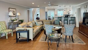 A spacious living room with a leather sofa, a glass coffee table, and a dining area with green chairs. A kitchen with white cabinets and a TV is visible in the background. - Oceanfront w/ sandy beach all day! 4100sq ft. Retreat w/ Private walk, Pet’s ok! (Fripp Island)
