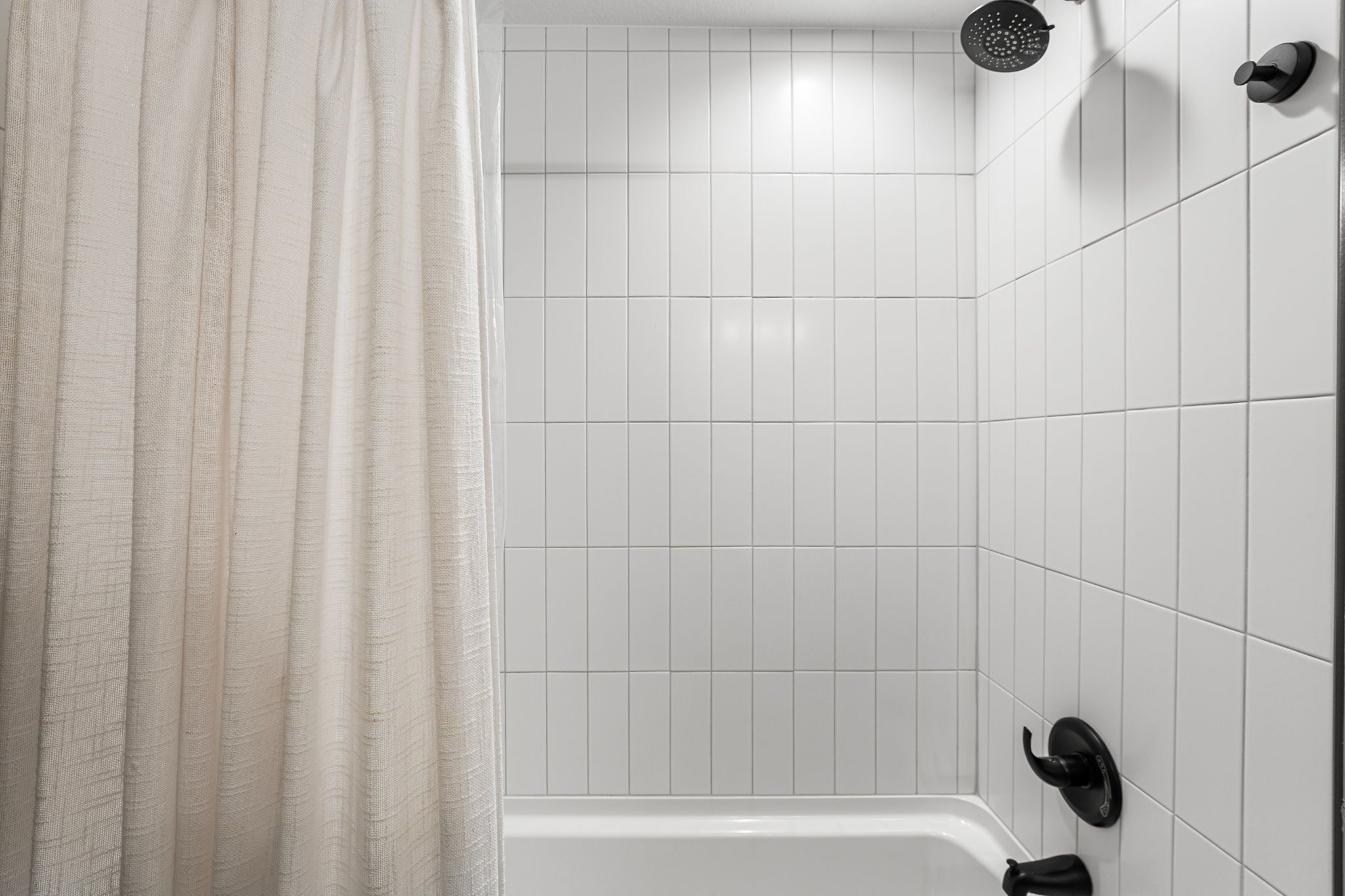 Une salle de bain avec des murs carrelés de carreaux blancs, une douche dotée d’un pommeau de douche noir et d’un rideau blanc.