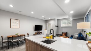 A modern kitchen with a central island, a dining area with chairs, a flat-screen TV, and a window with blinds.