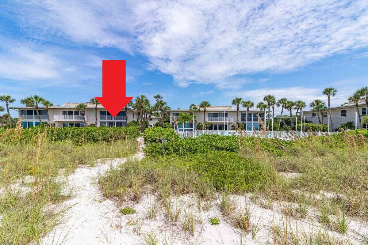 Un hotel frente a la playa con un camino que lleva a la entrada, rodeado de arena y vegetación.