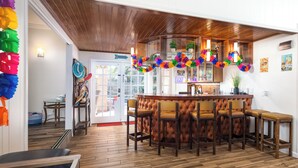 Zona de bar con mostrador de madera y varios taburetes. Una guirnalda colorida cuelga sobre la barra.