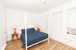 Ein Schlafzimmer mit einem Himmelbett, weißen Wänden, Holzboden und einem Fenster mit Jalousien.