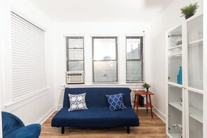 Ein Zimmer mit einem blauen Sofa, zwei gemusterten Kissen, einem kleinen Holznachttisch und einer weißen Regaleinheit.