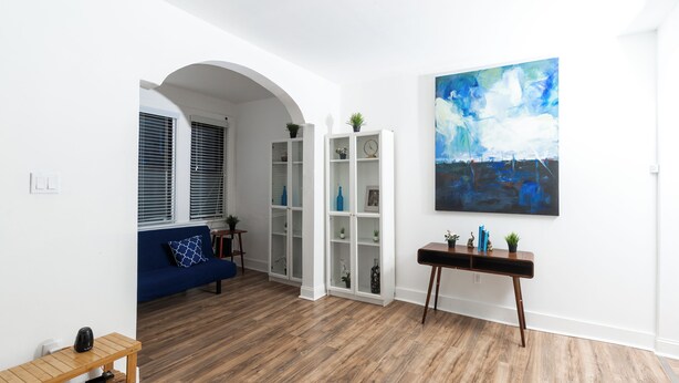 Un salón moderno con un sofá azul, suelo de madera, paredes blancas y un cuadro abstracto grande.