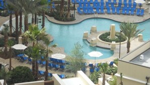 Área de piscina en un resort con sillas reclinables azules y sombrillas, rodeada de palmeras.
