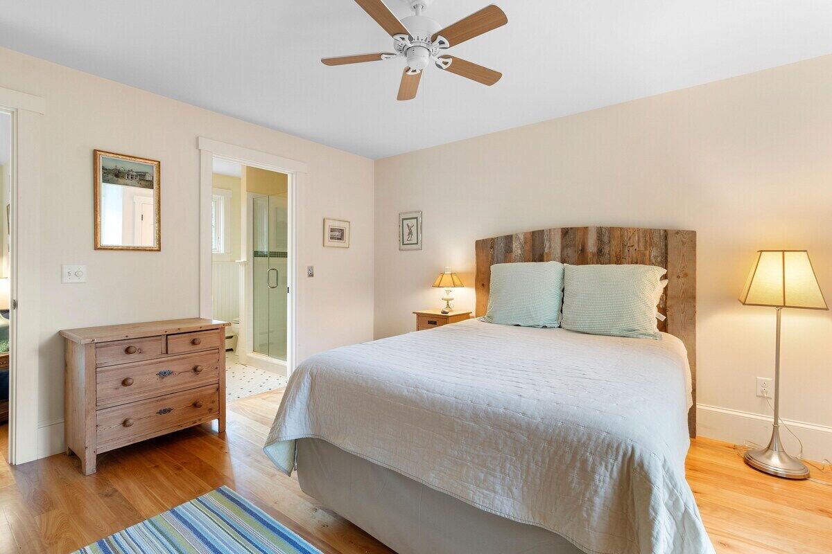 A bedroom with a bed, a dresser, a ceiling fan, a lamp, and a door leading to another room.