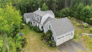 Une grande maison à deux étages avec un garage, entourée d’une forêt dense.