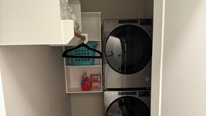 Une buanderie équipée de deux machines à laver à chargement frontal superposées, d’une étagère contenant de la lessive et d’un panier à linge suspendu.