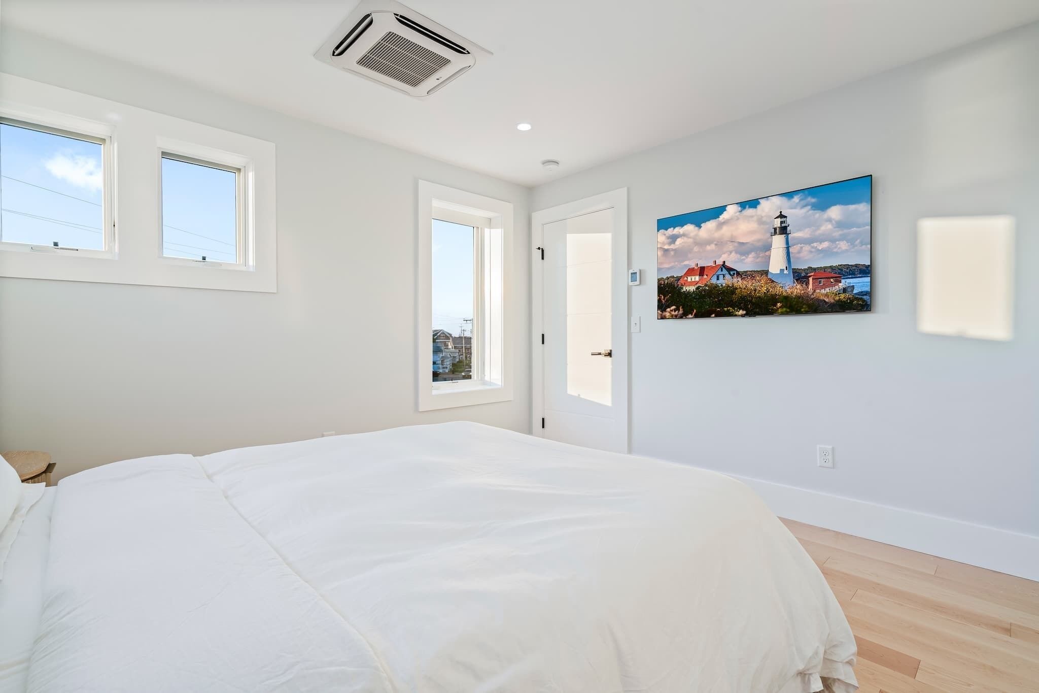 Ein Schlafzimmer mit einem großen Bett, zwei Fenstern, einer Tür und einem Gemälde von einem Leuchtturm.