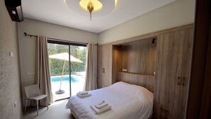 A bedroom with a large bed, a chair, a wooden wardrobe, and a view of an outdoor swimming pool.