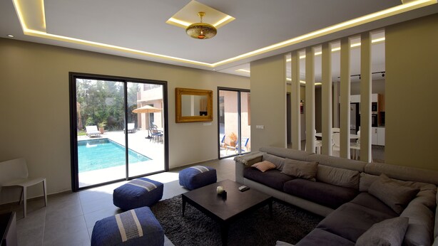 A modern living room with a sectional sofa, ottomans, a coffee table, and a large window overlooking a pool.