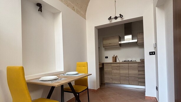 A dining area with a table and two yellow chairs, a kitchen in the background, and a tiled floor. - Casaggiù traditional house just steps from the towncenter & seaside destinations (San Vito dei Normanni)